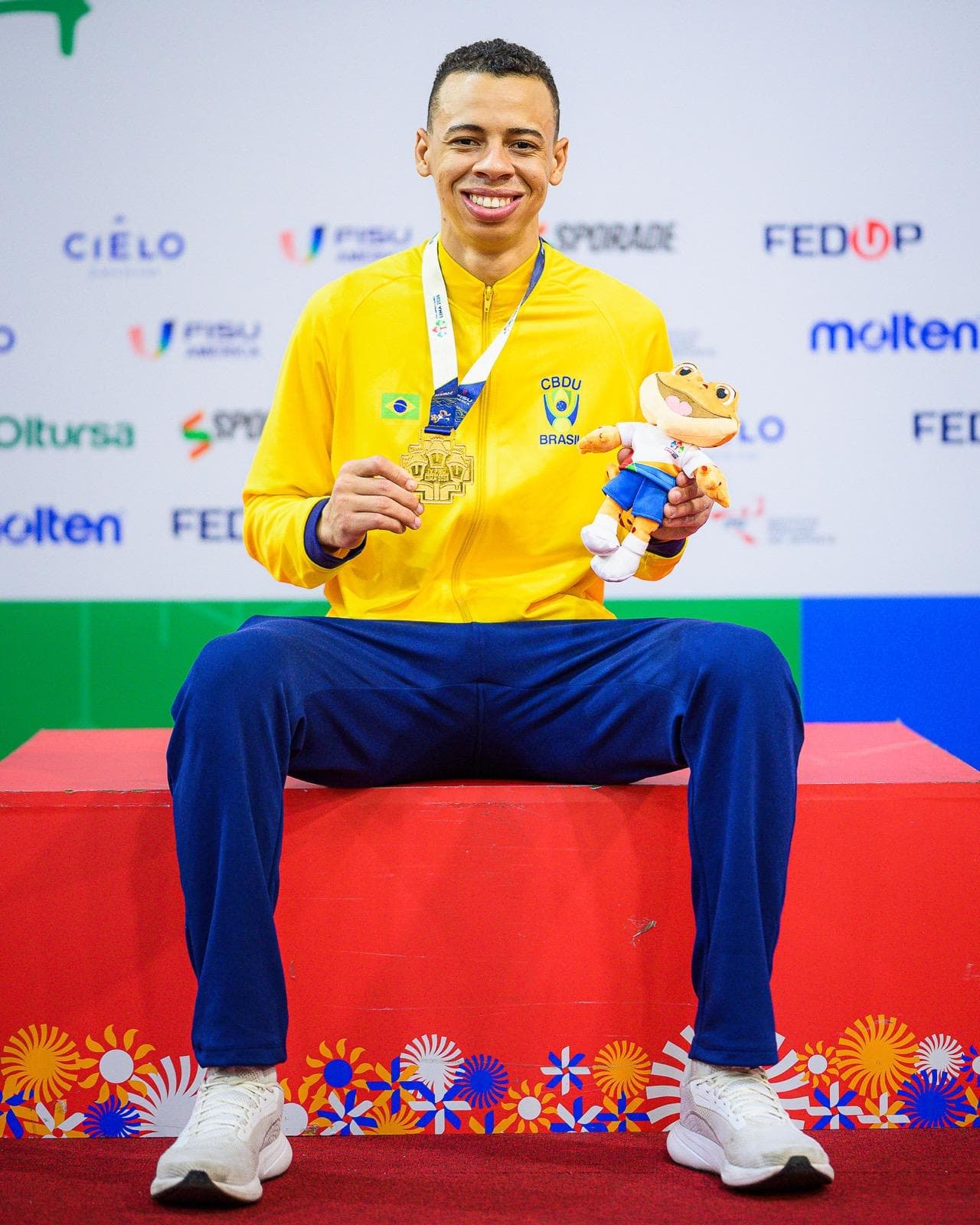 Luiz Augusto sentado no pódio com a medalha de ouro e o mascote da competição
